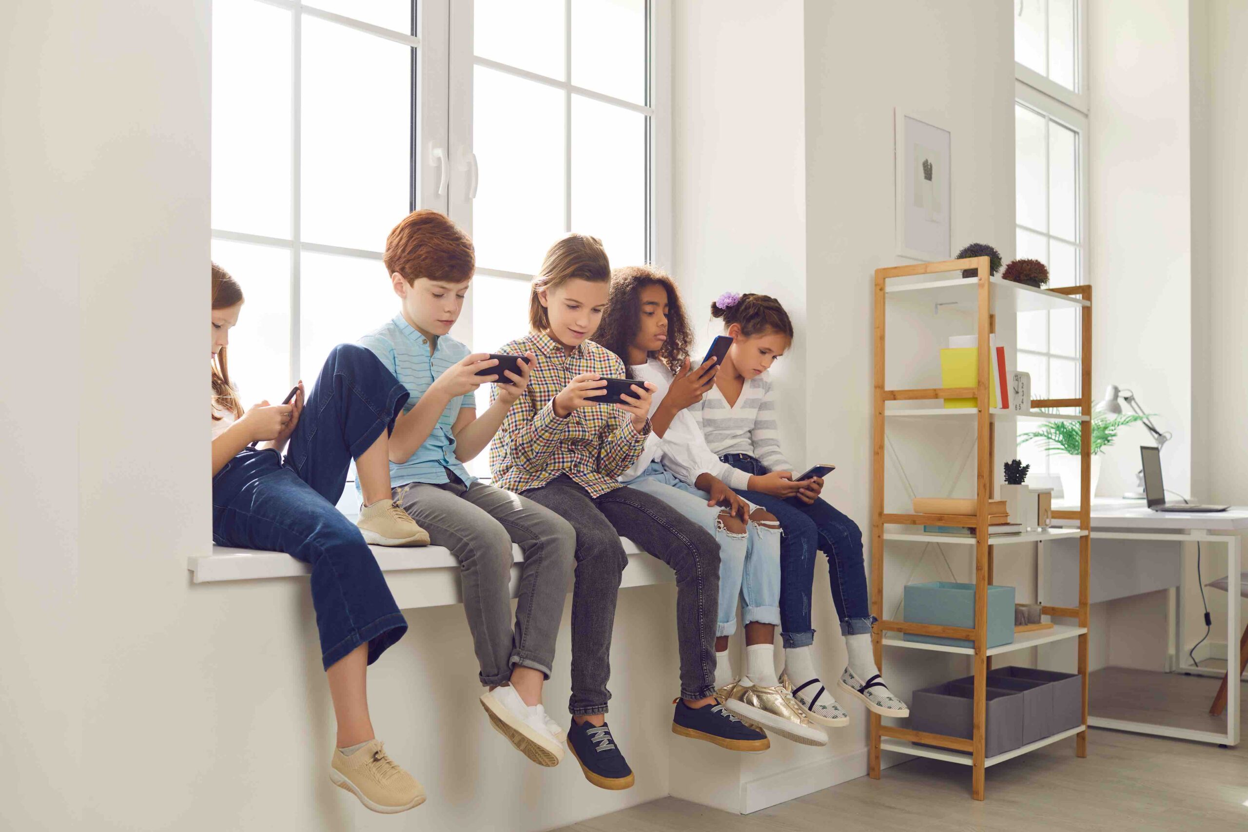 Diverse group of 8-10 year old children sitting on windowsill, playing online phone games and ignoring each other. Concept of gadget addiction and excessive use of social media and mobile devices